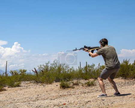 Photo for A man with earmuffs aiming an AK-47 automatic rifle with a scope on a sunny day in the Arizona Summer Dessert - Royalty Free Image