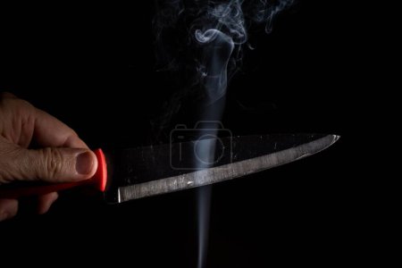 Téléchargez les photos : Une main masculine tenant un couteau d'argent pointu avec de la fumée sur fond noir - en image libre de droit