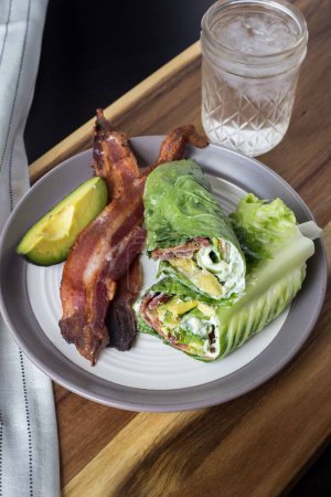 Photo for A vertical closeup shot of the lettuce wraps with bacon and avocado on a gray plate with a glass of iced water on a wooden table - Royalty Free Image