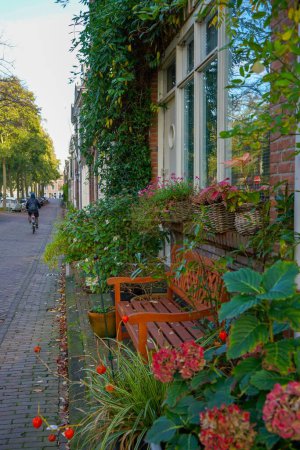 Foto de Un banco de madera rodeado de flores y hojas en frente de una casa de ladrillo en una calle empedrada en Amsterdam, Países Bajos - Imagen libre de derechos