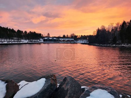 Téléchargez les photos : Un paysage pittoresque d'un lac entouré de forêts et de maisons lors d'un coucher de soleil coloré un jour d'hiver - en image libre de droit
