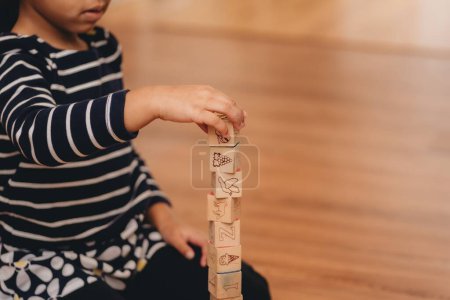 Photo for A preschool girl, playing with wooden letter blocks at home on the hardwood floor, the concept of early education - Royalty Free Image