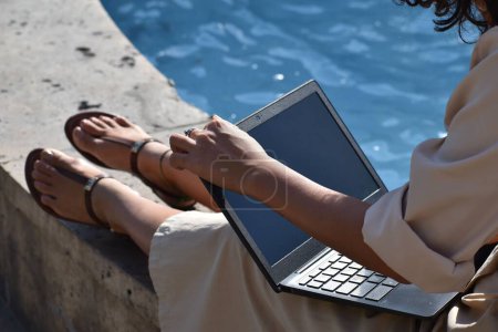 Photo for A woman sitting on a ledge and using a laptop at the sunlight - Royalty Free Image