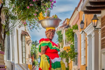 Foto de Un metraje regular de una Palenquera sosteniendo un gran tazón de frutas en la cabeza, vestida con ropa colorida en una calle de Cartagena, España - Imagen libre de derechos