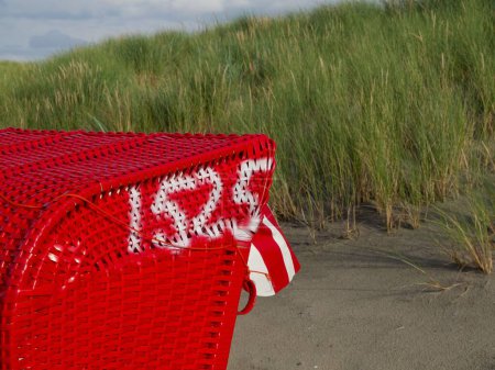 Téléchargez les photos : Gros plan d'une chaise de plage à capuchon rouge sur la plage de sable de l'île Juist, par une journée nuageuse, avec de l'herbe verte en arrière-plan - en image libre de droit