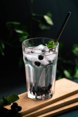 Photo for A vertical shot of a lemonade with ice and blackberry on a wooden board on the blurred background - Royalty Free Image