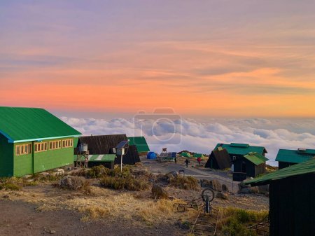 Téléchargez les photos : Les cabanes Horombo sur le mont Kilimandjaro avec une mer de nuages pelucheux autour d'eux sous un ciel coloré - en image libre de droit