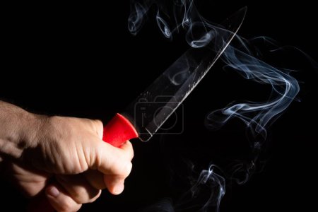 Téléchargez les photos : Une main masculine tenant un couteau d'argent pointu avec de la fumée sur fond noir - en image libre de droit