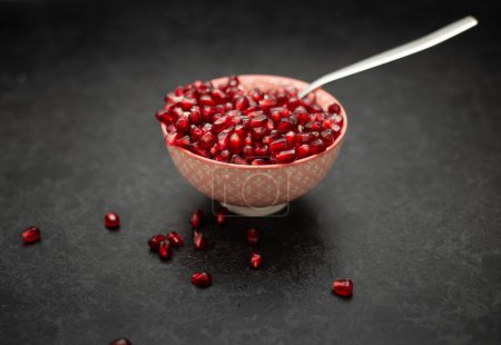 Foto de Un primer plano de un tazón lleno de semillas jugosas de granada roja, servido con una cuchara, y semillas de granada alrededor del tazón, sobre un fondo gris - Imagen libre de derechos