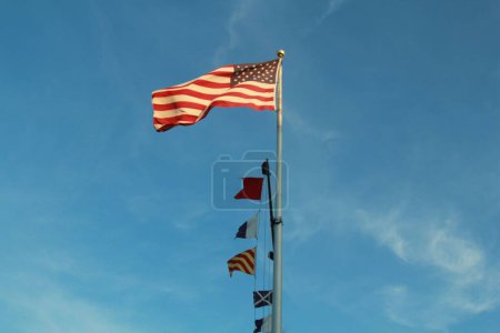 Foto de La bandera de EE.UU. en un poste metálico alto con otras banderas junto a ella bajo el cielo azul - Imagen libre de derechos