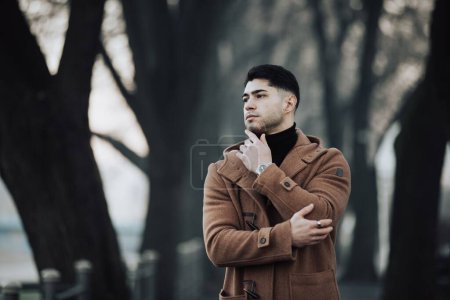 Téléchargez les photos : Un jeune homme à la mode en manteau d'hiver chic posant à côté d'un arbre - en image libre de droit