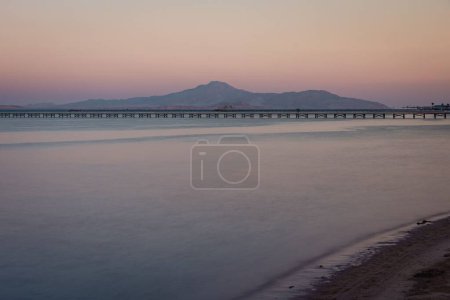 Téléchargez les photos : La mer calme et la plage de sable avec un quai en bois et des montagnes contre le coucher de soleil rose - en image libre de droit
