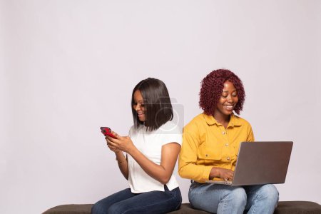 Photo for Two black ladies sitting together using laptop and phone - Royalty Free Image