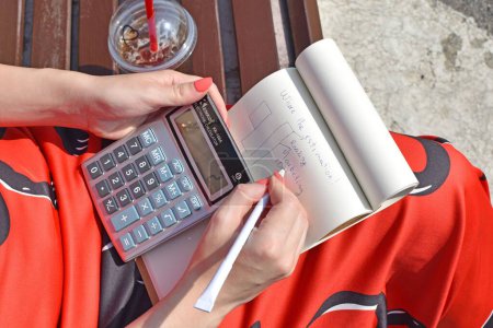 Téléchargez les photos : Gros plan d'une femme assise sur un banc avec un verre et écrivant sur un carnet avec une calculatrice - en image libre de droit