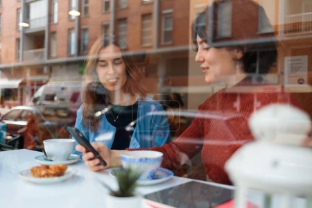 Photo for The two women visible through window glass sitting in the cafe, drinking coffee and chatting - Royalty Free Image