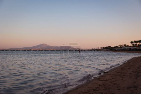 Foto de El mar tranquilo y la playa de arena con un muelle de madera y montañas contra la puesta de sol rosa - Imagen libre de derechos