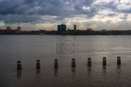 Téléchargez les photos : Une vue horizontale du New Jersey à travers le fleuve Hudson avec un ciel orageux et des pylônes de jetée avec un reflet dans l'eau - en image libre de droit