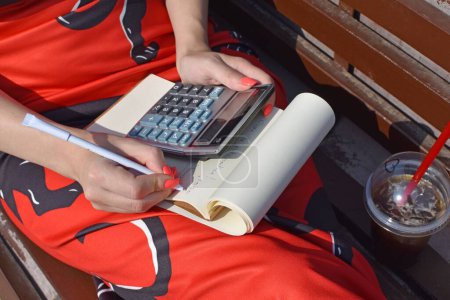 Foto de Un primer plano de una mujer sentada en el banco con una bebida y escribiendo en el cuaderno con el uso de la calculadora - Imagen libre de derechos
