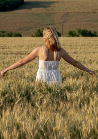 Foto de Un disparo vertical de una joven con un vestido blanco de pie en el campo de cebada durante la hora dorada - Imagen libre de derechos