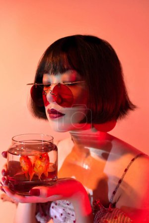 Téléchargez les photos : Un portrait d'une femme sexy attrayante d'Asie de l'Est avec des lunettes de soleil rouges tenant des poissons rouges dans un verre et posant - en image libre de droit