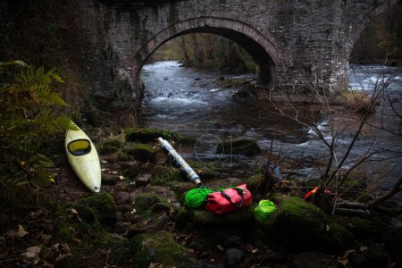 Photo for A kayak boat and camping tools on a river bank with ancient stone arch bridge in Ebbw Vale, Wales - Royalty Free Image