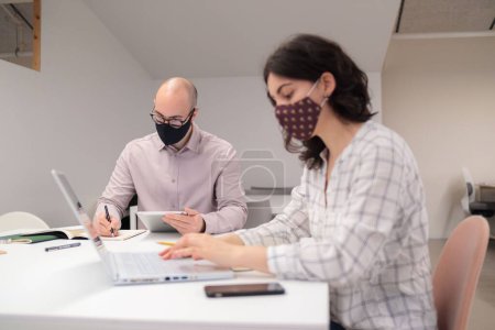 Photo for The young colleagues in protective face masks sitting by the table with documents and laptop at office - Royalty Free Image