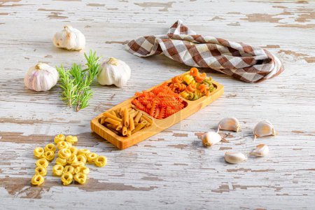 Foto de Un primer plano de diferentes tipos de pasta italiana, macarrones presentados en una tabla de madera junto a un poco de ajo - Imagen libre de derechos