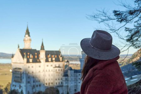 Téléchargez les photos : Une touriste avec un chapeau gris admirant le château de Neuschwanstein en Allemagne depuis une colline - en image libre de droit