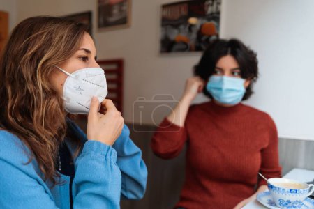 Téléchargez les photos : Les deux femmes portant des masques médicaux, assises au café, buvant du café et bavardant - en image libre de droit