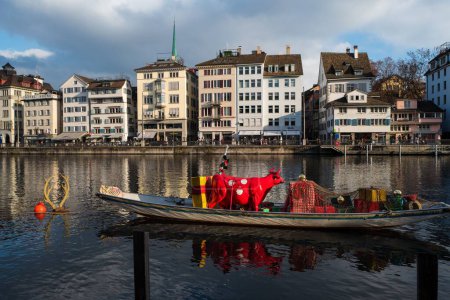 Foto de Una vista horizontal del río Limmat de Zurich con un bote flotante, una vaca roja dentro y edificios antiguos a lo largo del río - Imagen libre de derechos