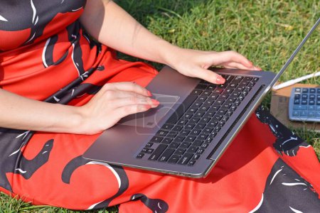 Foto de Un primer plano de una mujer sentada en la hierba y el uso de la computadora portátil y hay calculadora con pluma al lado de su pierna - Imagen libre de derechos
