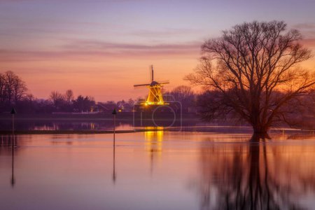 Photo for A scenic view of a calm waters with luminated old windmill in the background - Royalty Free Image