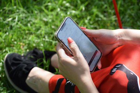 Téléchargez les photos : Un gros plan d'une fille cherchant quelque chose sur son téléphone, iPhone tout en étant assis sur l'herbe dans un parc - en image libre de droit