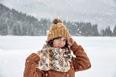 Photo for A woman in a cozy winter sweater and scarf looking at the camera under falling snow in the wilderness - Royalty Free Image