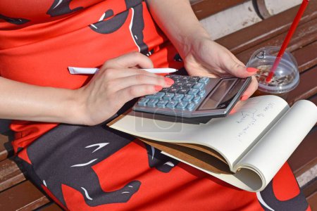 Téléchargez les photos : Gros plan d'une femme assise sur un banc avec un verre, à l'aide d'une calculatrice pour écrire sur un cahier - en image libre de droit