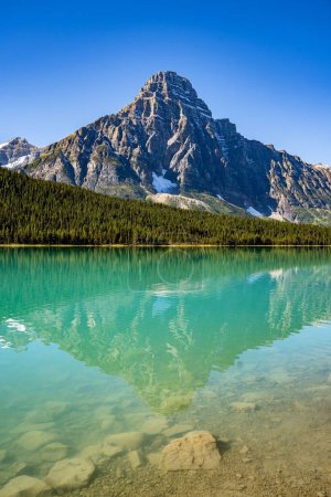 Foto de Una magnífica vista de la superficie de un lago de montaña transparente, con un bosque siempreverde y laderas nevadas en el fondo, en un día soleado - Imagen libre de derechos