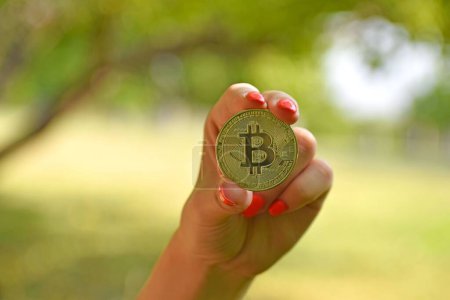 Foto de La mano de una mujer sosteniendo una moneda de oro bitcoin sobre un fondo borroso natural - concepto de criptomoneda - Imagen libre de derechos