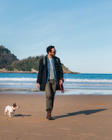Téléchargez les photos : Un jeune homme blanc beau portant des lunettes de soleil avec son chien sur la plage par une journée ensoleillée - en image libre de droit