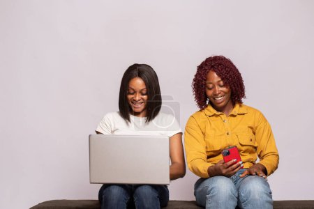 Foto de Dos chicas negras sentadas juntas usando teléfono y portátil - Imagen libre de derechos