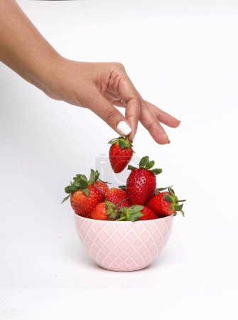 Téléchargez les photos : Plan vertical d'une main cueillant des fraises dans un bol rose sur une surface blanche - en image libre de droit