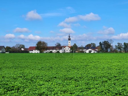 Téléchargez les photos : Un grand champ vert et l'église paroissiale de Saint-Vitus sur une distance à Iffeldorf, Allemagne avec un ciel bleu - en image libre de droit