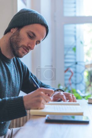 Téléchargez les photos : Un jeune homme écrit dans un cahier près de la fenêtre sur une table en bois - en image libre de droit