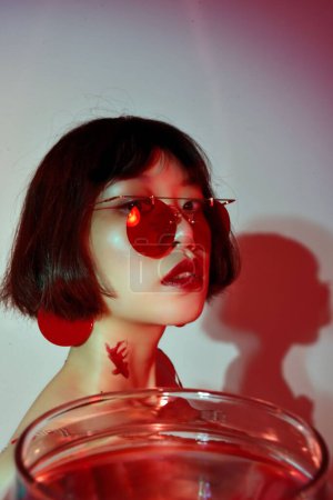 Foto de Un retrato de una atractiva mujer sexy de Asia Oriental con gafas de sol rojas sosteniendo peces de colores en un vaso y posando - Imagen libre de derechos
