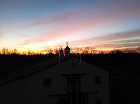 Téléchargez les photos : Une maison individuelle et la silhouette des girouettes et des arbres à cheval contre le ciel orange-rouge au coucher du soleil - en image libre de droit
