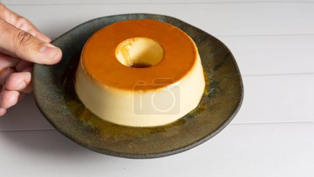 Photo for A closeup shot of a man holding a Spanish and Latin American traditional pudding dessert, Flan, served on a black plate on a white wooden table - Royalty Free Image