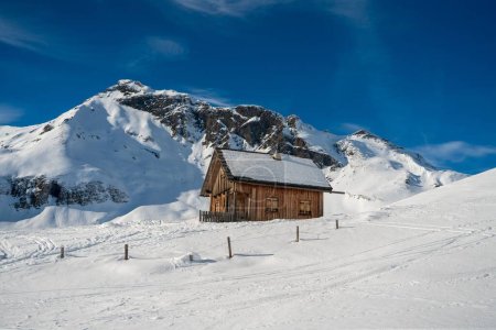 Foto de Una pequeña cabaña de madera en un frío día de invierno soleado rodeado de las montañas nevadas - Imagen libre de derechos