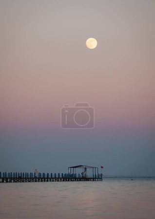 Foto de Un plano vertical del mar tranquilo y el muelle de madera contra el cielo rosado del atardecer con una luna llena - Imagen libre de derechos