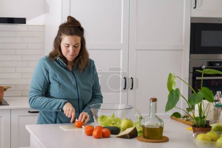 Photo for Beautiful young long-haired brunette girl cutting fresh tomatoes to prepare a salad in the kitchen - Royalty Free Image