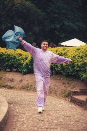 Téléchargez les photos : Plan vertical d'un homme hispanique heureux marchant dans un parc avec des ballons bleus - en image libre de droit