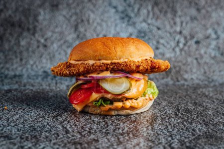 Téléchargez les photos : Gros plan d'un grand cheeseburger au poulet frit avec des légumes sur une surface grise - en image libre de droit